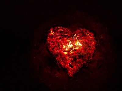 A glowing heart shape formed by embers and flames against a dark background. The heart is outlined in bright red and orange colors, giving it a vibrant and intense appearance.
