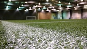 An indoor sports facility with artificial turf and overhead lighting. The focus is on the white boundary line on the field, and a soccer goal is visible in the distance. The lighting creates a well-illuminated environment with shadows cast on the turf.