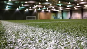 An indoor sports facility with artificial turf and overhead lighting. The focus is on the white boundary line on the field, and a soccer goal is visible in the distance. The lighting creates a well-illuminated environment with shadows cast on the turf.