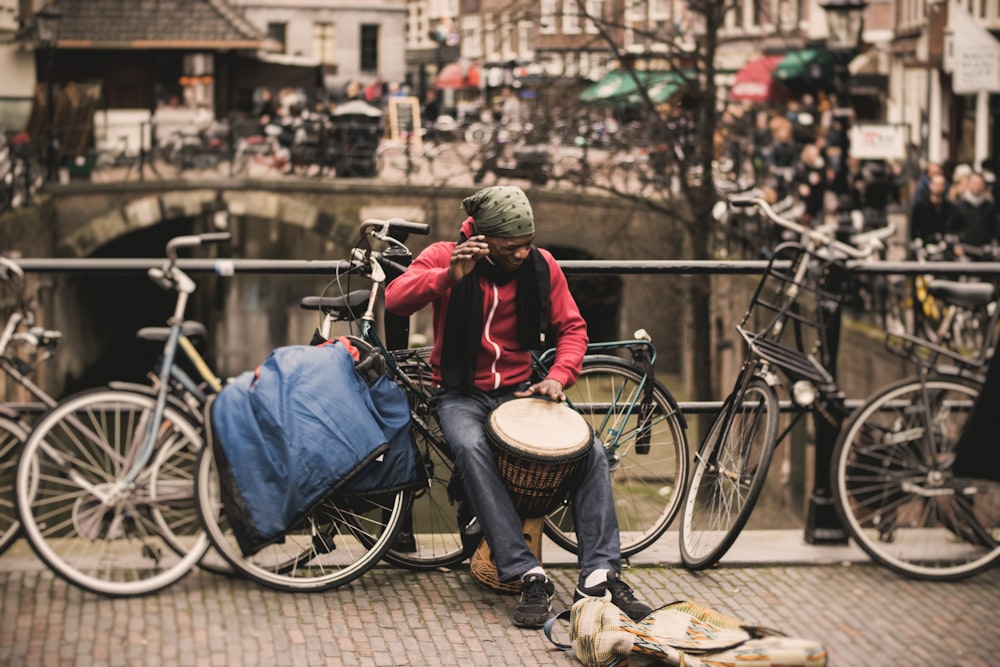 uomo in camicia rossa che suona il tamburo darbuka mentre è seduto sulla bici grigia vicino alla ringhiera del ponte