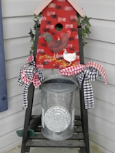 A decorative piece with a chicken theme is displayed on a porch. It features a red birdhouse painted with images of a rooster and a chicken. The birdhouse sits on a metal container, adorned with checkered black and red ribbons tied in bows. There is greenery, likely ivy, peeking out from behind the birdhouse.
