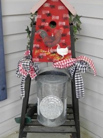 A decorative piece with a chicken theme is displayed on a porch. It features a red birdhouse painted with images of a rooster and a chicken. The birdhouse sits on a metal container, adorned with checkered black and red ribbons tied in bows. There is greenery, likely ivy, peeking out from behind the birdhouse.