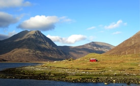 A vivid landscape features towering mountains under a clear blue sky with white clouds. In the foreground, a body of water flows alongside a grassy and rocky area. A distinct red van is parked on a grassy hill, adding a splash of color to the natural scene.