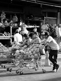 A busy market scene with people interacting and shopping. Several shopping carts are filled with a variety of fruits, including watermelons and pineapples. In the background, fresh produce is displayed on shelves, and a hanging scale is visible.