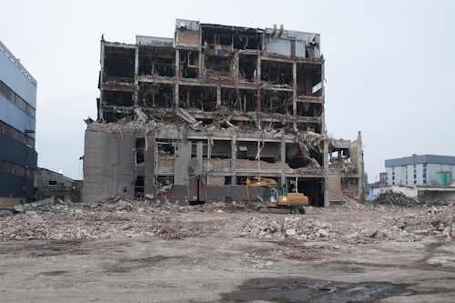 A multi-story building is in a state of partial demolition, with exposed floors and walls showing structural damage. There is debris scattered around, and a construction excavator is visible in the foreground. The sky is overcast, lending a somber atmosphere to the scene.