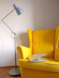 A bright yellow armchair with buttons on the backrest is positioned next to a tall, gray adjustable floor lamp. A modern silver laptop is open and placed on the seat of the chair. The room features wooden flooring and a plain, light-colored wall.