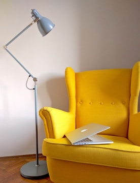 A bright yellow armchair with buttons on the backrest is positioned next to a tall, gray adjustable floor lamp. A modern silver laptop is open and placed on the seat of the chair. The room features wooden flooring and a plain, light-colored wall.