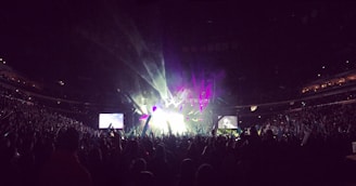 A large, packed concert arena with an enthusiastic crowd. Bright stage lights and colorful lasers create an energetic atmosphere. Two large screens flank the stage, projecting live performances.