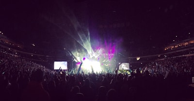 A large, packed concert arena with an enthusiastic crowd. Bright stage lights and colorful lasers create an energetic atmosphere. Two large screens flank the stage, projecting live performances.