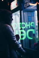 A person is standing next to a glass-fronted Prize Cube machine, with neon lettering reflecting on the glass. They are wearing a beanie and a jacket, holding something in their hand. The scene is dimly lit, imparting a moody atmosphere with visible neon green and blue lights.