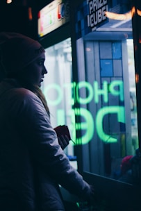 A person is standing next to a glass-fronted Prize Cube machine, with neon lettering reflecting on the glass. They are wearing a beanie and a jacket, holding something in their hand. The scene is dimly lit, imparting a moody atmosphere with visible neon green and blue lights.
