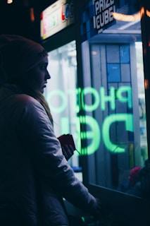 A person is standing next to a glass-fronted Prize Cube machine, with neon lettering reflecting on the glass. They are wearing a beanie and a jacket, holding something in their hand. The scene is dimly lit, imparting a moody atmosphere with visible neon green and blue lights.