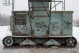 An old, rusted industrial machine with visible tracks and side panels covered in graffiti. The device appears to be abandoned in an outdoor setting with a foggy, leafless tree line in the background. Various cables and metal components are visible, adding to the aged and neglected appearance.