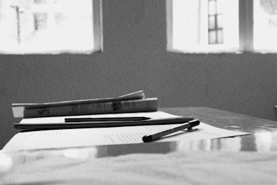 A black and white photograph featuring a close-up of a table with various objects on it. There are two pens placed on top of a few sheets of paper, and a closed book lies on the table next to the papers. The background reveals a blurred view of three window frames with light streaming in.