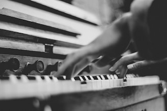 Black and white photography of a person's hands playing a piano or an organ. The keys and part of the instrument are visible, creating a focus on the act of playing music.