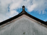 A traditional architectural roof with ornate patterns exists against a backdrop of a partly cloudy sky. The roof features a decorative edge with subtle red and gold detailing, and the tip slightly curves upward.