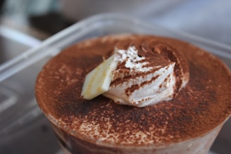 A close-up of a dessert featuring a fluffy whipped cream topping dusted with cocoa powder. The dessert appears to be in a transparent container, emphasizing the rich, creamy textures.