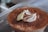 A close-up of a dessert featuring a fluffy whipped cream topping dusted with cocoa powder. The dessert appears to be in a transparent container, emphasizing the rich, creamy textures.