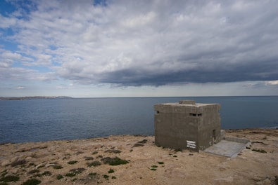 A concrete bunker stands on a rocky coastline under a cloudy sky. The sea is calm with waves gently lapping the shore. The landscape is sparse with patches of grass scattered on the rocky terrain.