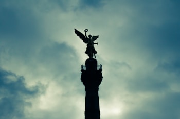 A statue of an angel with wings spread wide stands atop a tall column against a dramatic, cloudy sky. The silhouette of the angel is in sharp contrast to the soft, diffused light filtering through the clouds.