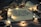 A notebook is open to a page with handwritten text surrounded by warm, glowing string lights. A ceramic mug lies overturned nearby, with additional handwriting visible on it. Dried flowers and scattered photographs are placed around, creating an intimate and cozy atmosphere.