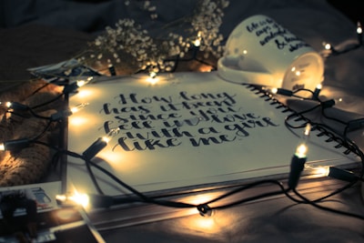 A notebook is open to a page with handwritten text surrounded by warm, glowing string lights. A ceramic mug lies overturned nearby, with additional handwriting visible on it. Dried flowers and scattered photographs are placed around, creating an intimate and cozy atmosphere.