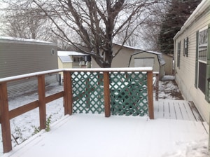 A snow-covered wooden deck with a lattice fence overlooks a backyard area. There are two structures with slanted roofs in the background, and a large tree stands behind them. The area around the deck is covered with a light layer of snow, and the branches of the trees are bare, indicating a winter season.