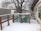 A snow-covered wooden deck with a lattice fence overlooks a backyard area. There are two structures with slanted roofs in the background, and a large tree stands behind them. The area around the deck is covered with a light layer of snow, and the branches of the trees are bare, indicating a winter season.