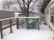 A snow-covered wooden deck with a lattice fence overlooks a backyard area. There are two structures with slanted roofs in the background, and a large tree stands behind them. The area around the deck is covered with a light layer of snow, and the branches of the trees are bare, indicating a winter season.