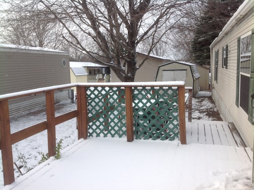 A snow-covered wooden deck with a lattice fence overlooks a backyard area. There are two structures with slanted roofs in the background, and a large tree stands behind them. The area around the deck is covered with a light layer of snow, and the branches of the trees are bare, indicating a winter season.