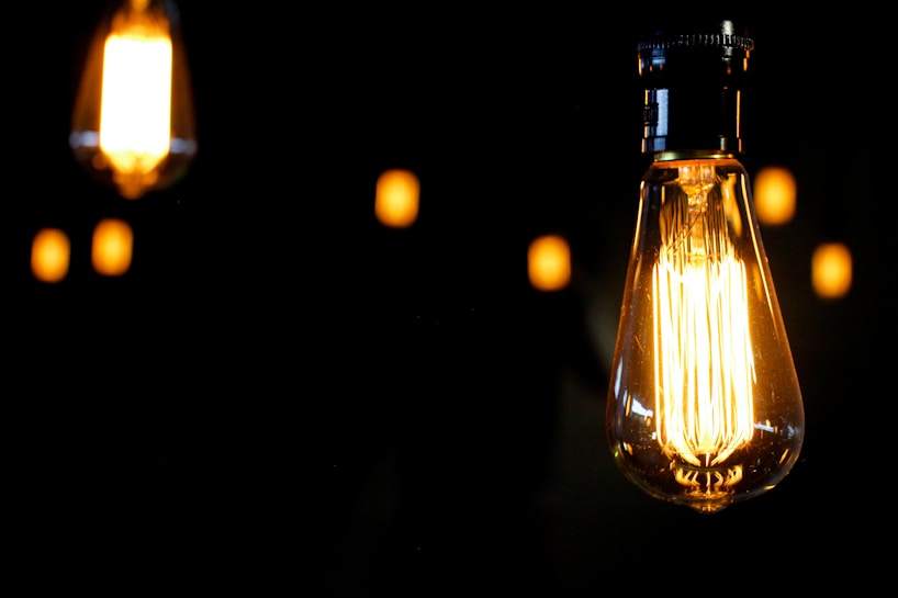 A close-up view of a glowing Edison-style light bulb against a dark background. The bulb emits a warm, amber glow with its filament visible. In the background, more out-of-focus bulbs are visible, contributing to a cozy and intimate atmosphere.