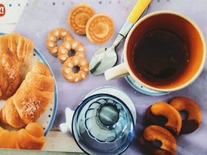 A breakfast setting with a warm cup of coffee, surrounded by a variety of pastries including croissants and different shaped cookies. The setup includes a reflective spoon and a stylized glass cup placed on a light purple tablecloth.