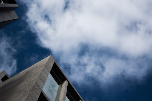 A modern building intersects with a sky filled with clouds. The structure features a geometric design with large window panels, contrasted against the backdrop of a partly cloudy blue sky.