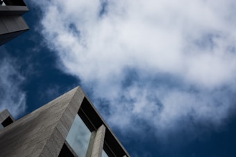 A modern building intersects with a sky filled with clouds. The structure features a geometric design with large window panels, contrasted against the backdrop of a partly cloudy blue sky.