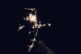 A hand is holding a brightly lit sparkler against a dark background. The sparkler emits glowing, sparkling light and creates a sense of celebration or festivity.