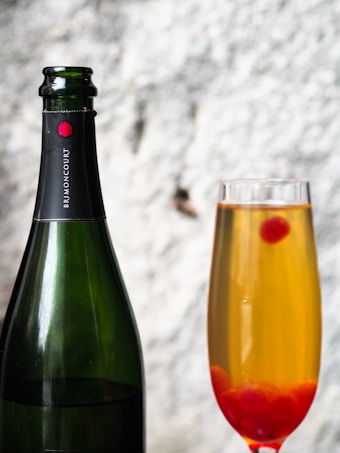 A black champagne bottle with a red emblem on the label stands next to a filled champagne flute containing a golden bubbly liquid and several red berries at the bottom. The background appears to be a blurred, light-colored surface.