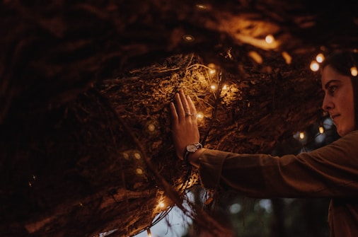 A person gently touches a textured, rough surface, possibly a tree branch or natural structure adorned with warm, glowing string lights. The setting is dimly lit, creating a cozy and intimate atmosphere. The person's expression is calm and contemplative.