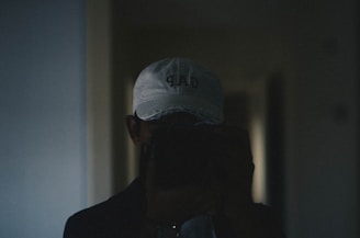 A person wearing a white cap with a logo on it is holding a camera or phone in front of their face, taking a photo. The image is dimly lit, giving it a moody atmosphere.