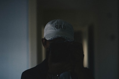 A person wearing a white cap with a logo on it is holding a camera or phone in front of their face, taking a photo. The image is dimly lit, giving it a moody atmosphere.
