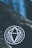 A black circular sticker with a white geometric diamond design placed on a rough, dark stone surface. The background features a blurred blue area, possibly indicating water or sky, with some small, scattered leaves.