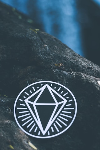 A black circular sticker with a white geometric diamond design placed on a rough, dark stone surface. The background features a blurred blue area, possibly indicating water or sky, with some small, scattered leaves.