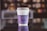 A single white disposable coffee cup sits on a reflective surface, creating a mirror image of the cup. The cup has a purple sleeve with white text that includes promotional and brand information. The background is blurred, giving the focus to the cup.