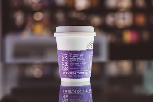 A single white disposable coffee cup sits on a reflective surface, creating a mirror image of the cup. The cup has a purple sleeve with white text that includes promotional and brand information. The background is blurred, giving the focus to the cup.