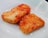 Two pieces of tofu are coated with a spicy red sauce and garnished with sesame seeds, placed on a white plate.