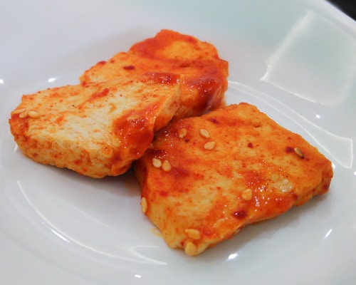 Two pieces of tofu are coated with a spicy red sauce and garnished with sesame seeds, placed on a white plate.