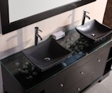 A modern bathroom vanity features two black, rectangular vessel sinks with sleek chrome faucets. Beneath the glass countertop, decorative stones are visible, adding an aesthetic element. A rolled-up towel rests between the sinks, which are situated on a dark wooden cabinet with multiple drawers and chrome handles.