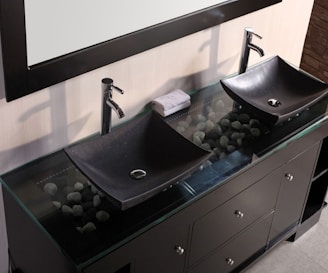 A modern bathroom vanity features two black, rectangular vessel sinks with sleek chrome faucets. Beneath the glass countertop, decorative stones are visible, adding an aesthetic element. A rolled-up towel rests between the sinks, which are situated on a dark wooden cabinet with multiple drawers and chrome handles.