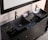 A modern bathroom vanity features two black, rectangular vessel sinks with sleek chrome faucets. Beneath the glass countertop, decorative stones are visible, adding an aesthetic element. A rolled-up towel rests between the sinks, which are situated on a dark wooden cabinet with multiple drawers and chrome handles.