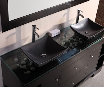 A modern bathroom vanity features two black, rectangular vessel sinks with sleek chrome faucets. Beneath the glass countertop, decorative stones are visible, adding an aesthetic element. A rolled-up towel rests between the sinks, which are situated on a dark wooden cabinet with multiple drawers and chrome handles.