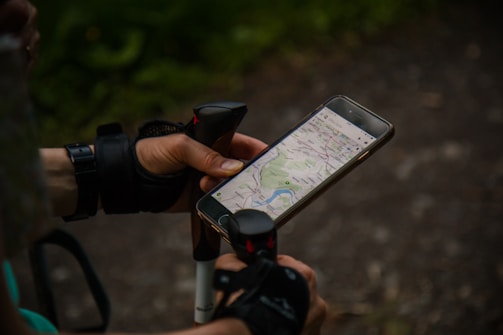 A person wearing fingerless gloves holds a smartphone displaying a map application. The person is also holding a walking pole, suggesting they might be hiking. The background is outdoors with a gravel path and greenery.